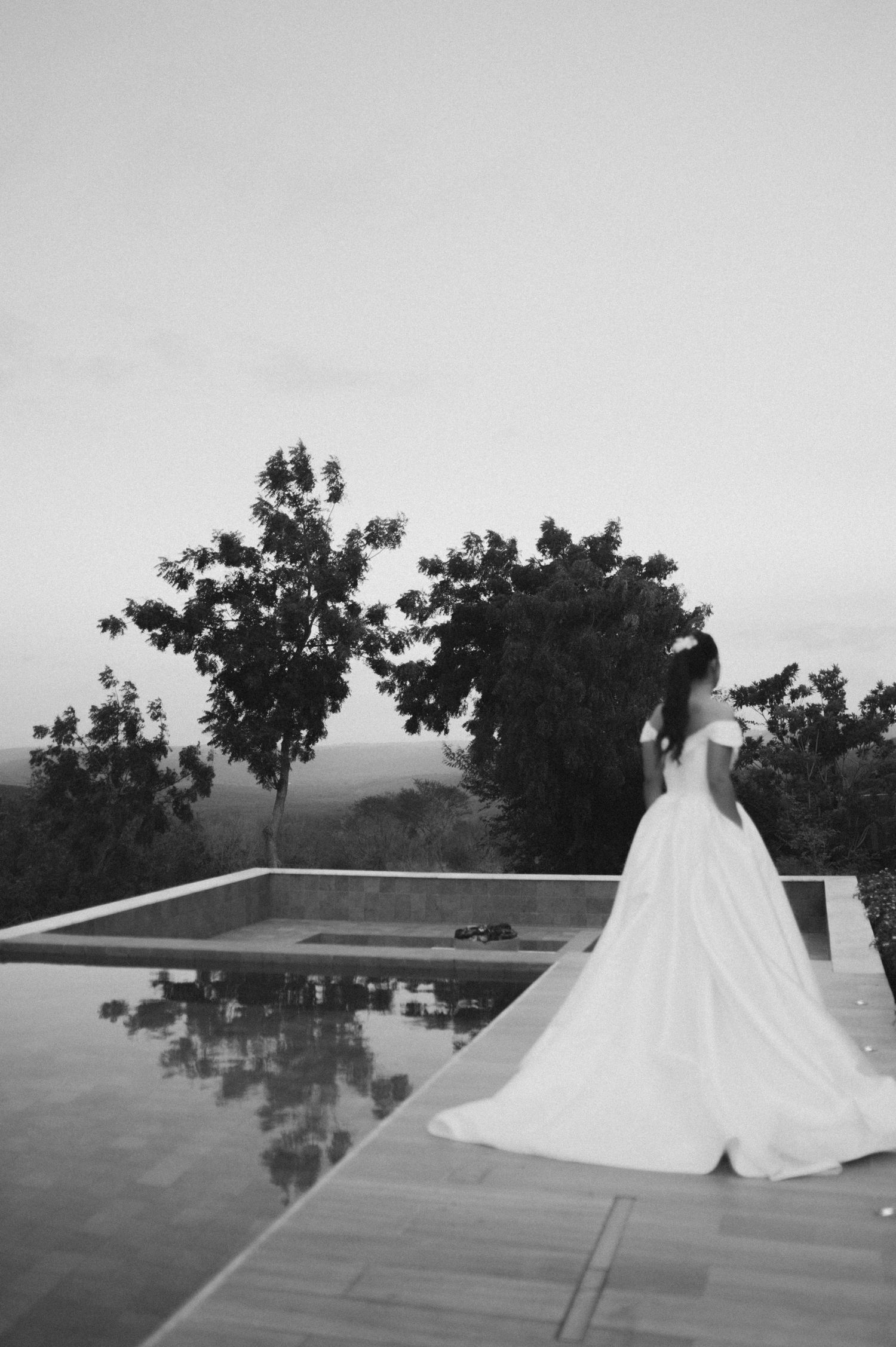 Elegant bride in a flowing white gown stands by a reflecting pool at dusk.
