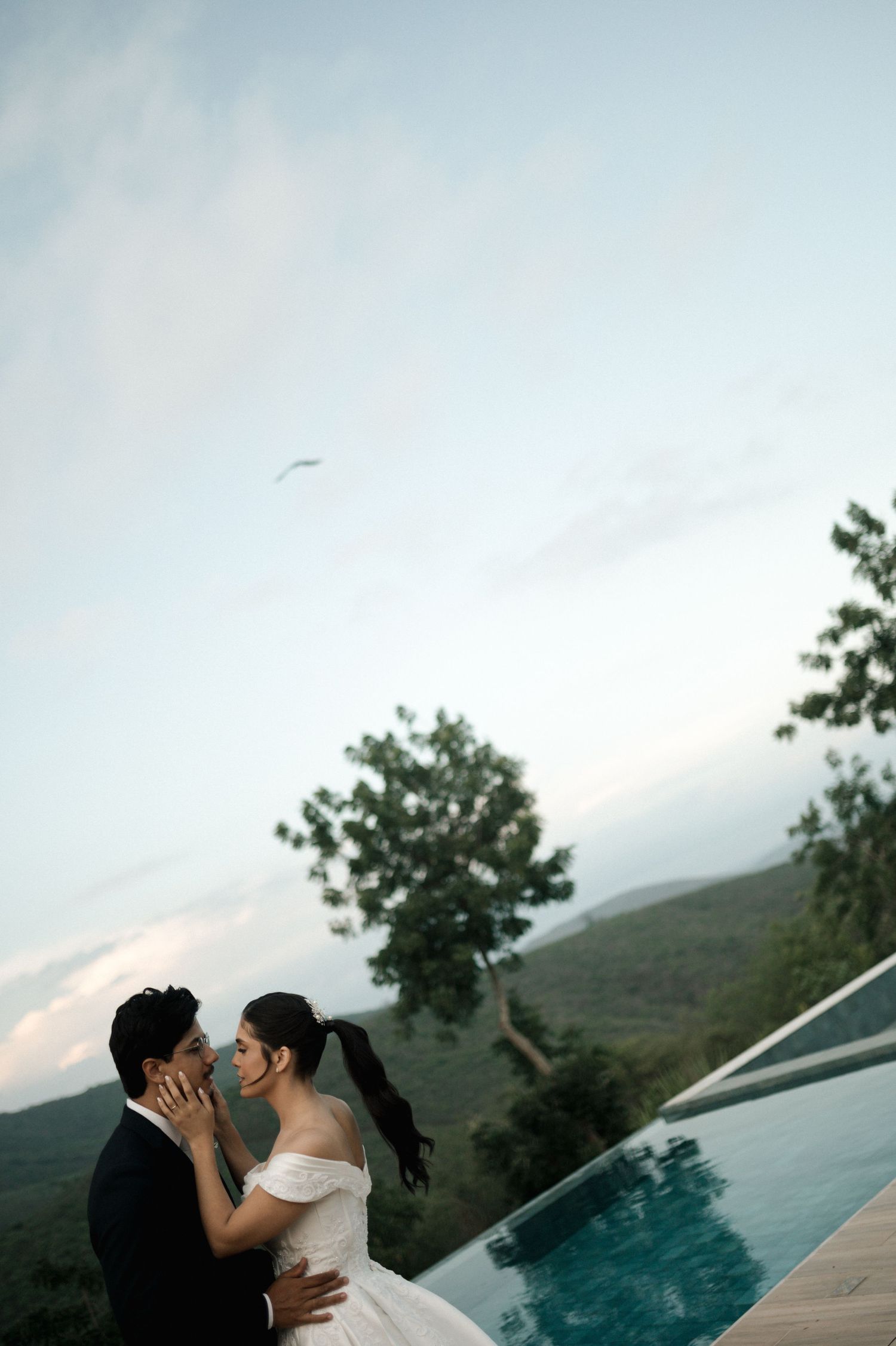 Bride and groom share a romantic moment by an infinity pool with lush mountain views at dusk.