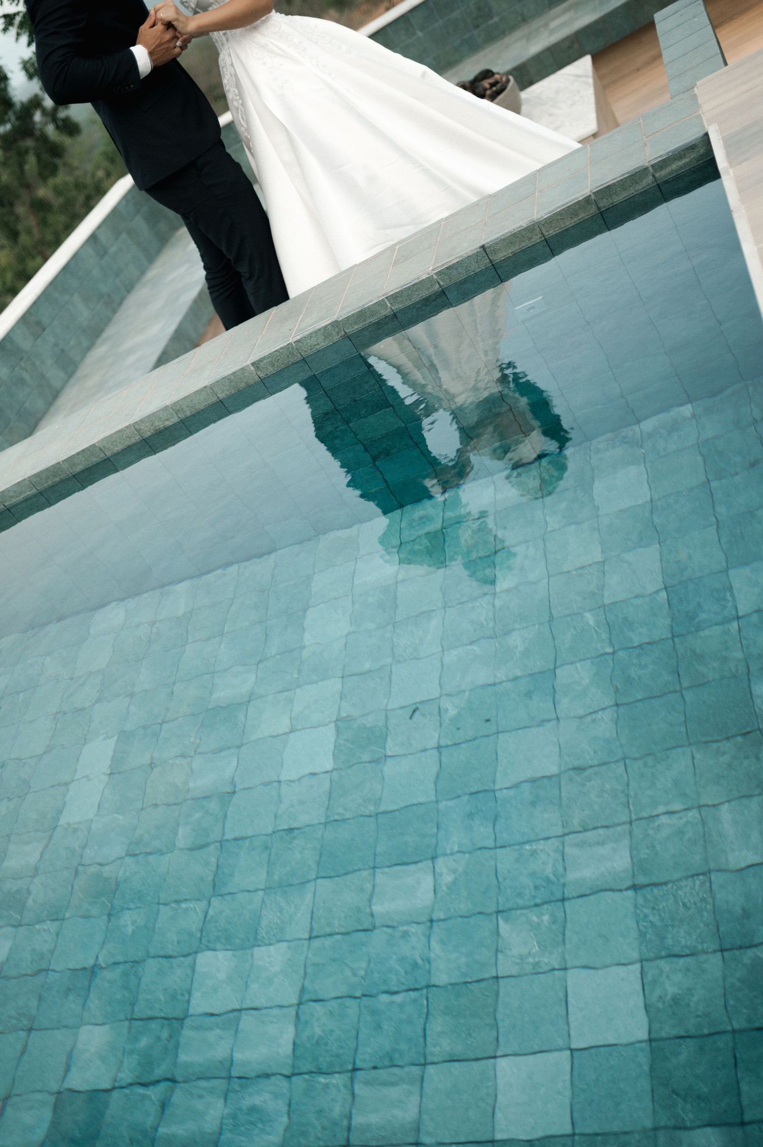 Bride and groom standing by a tiled pool, their reflection visible in the clear turquoise water on their wedding day.