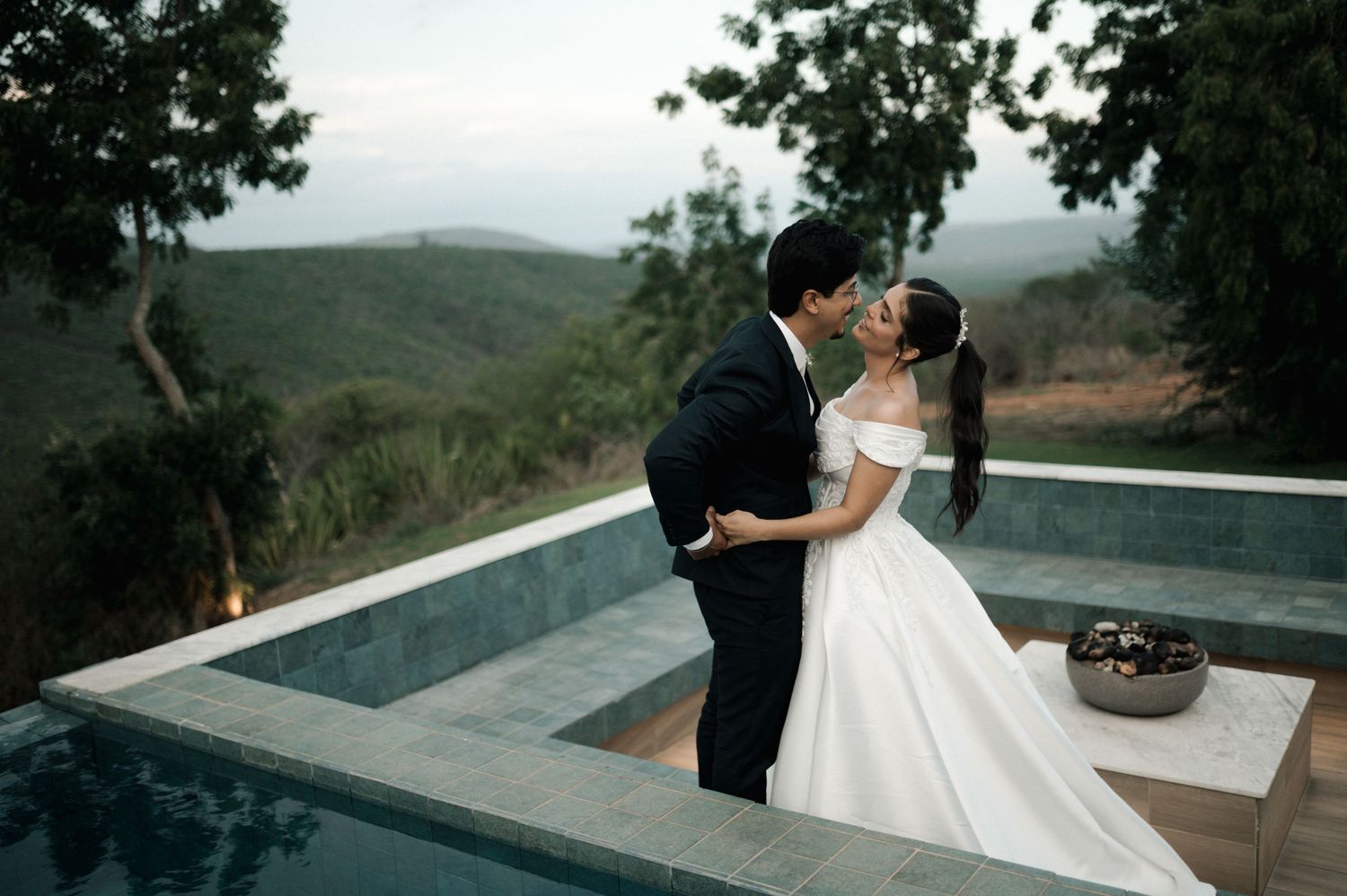 Bride and groom share a romantic kiss in elegant wedding attire beside an infinity pool with scenic mountain views.