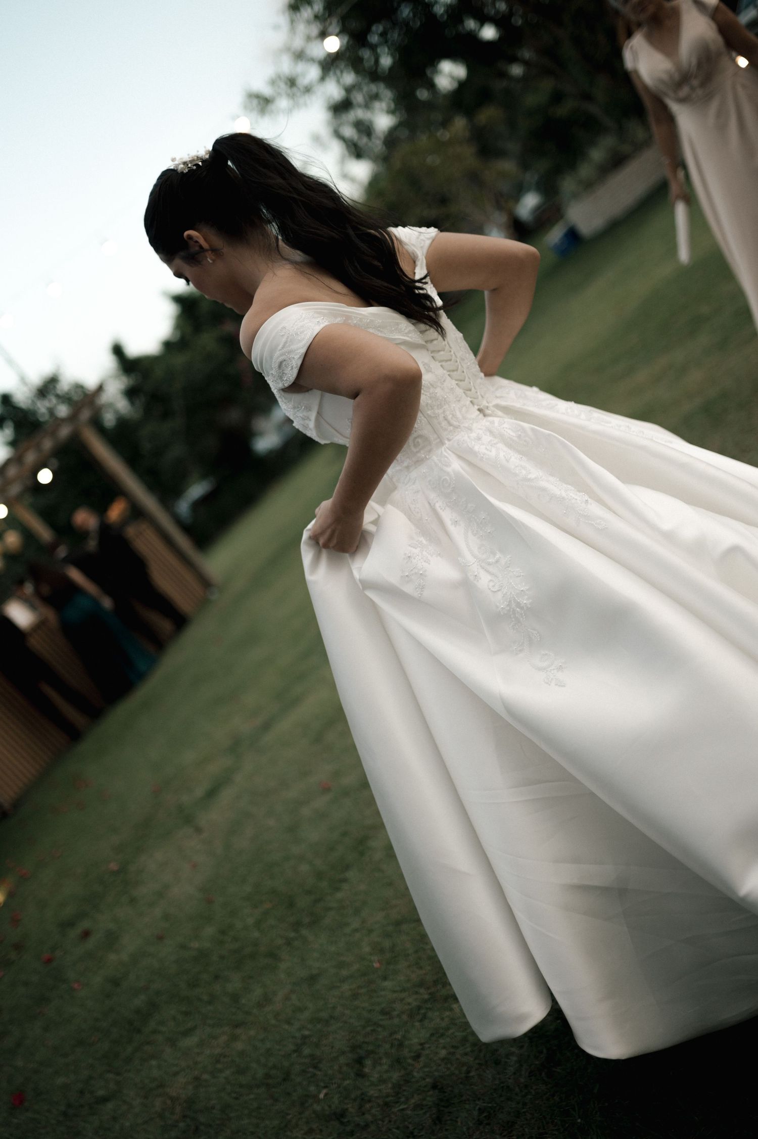 Bride in elegant off-shoulder white wedding gown twirling outdoors on green grass at a garden wedding ceremony.