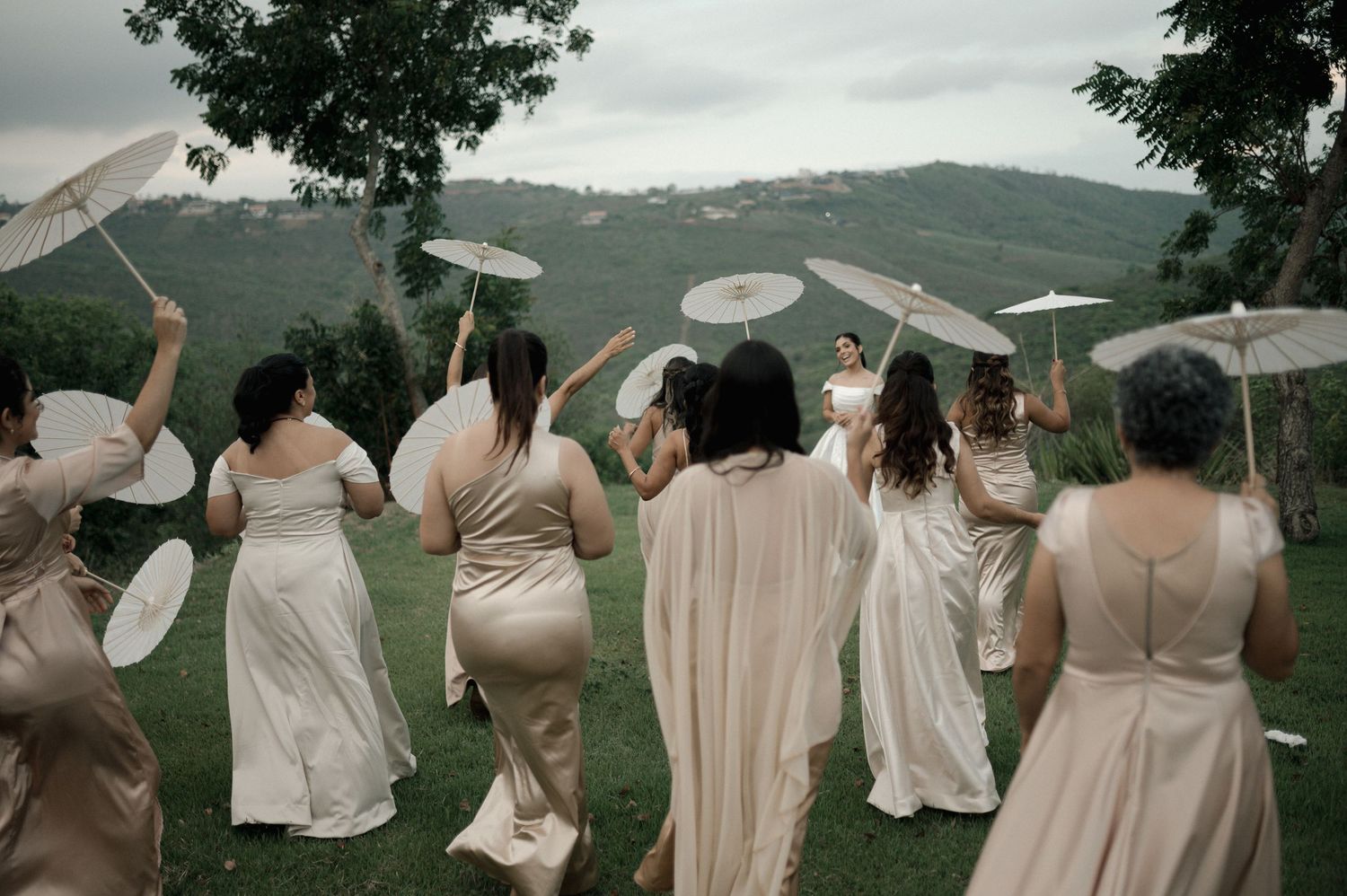 Bridesmaids in elegant gowns holding white parasols walk across lush green hillside at outdoor wedding ceremony.