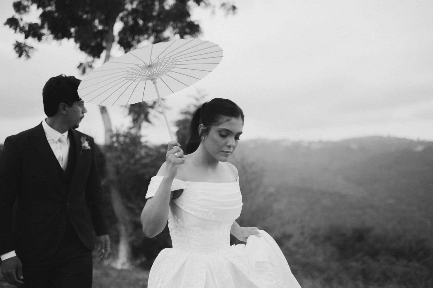 Bride in off-shoulder gown holding a white parasol with groom in black suit against scenic hills.