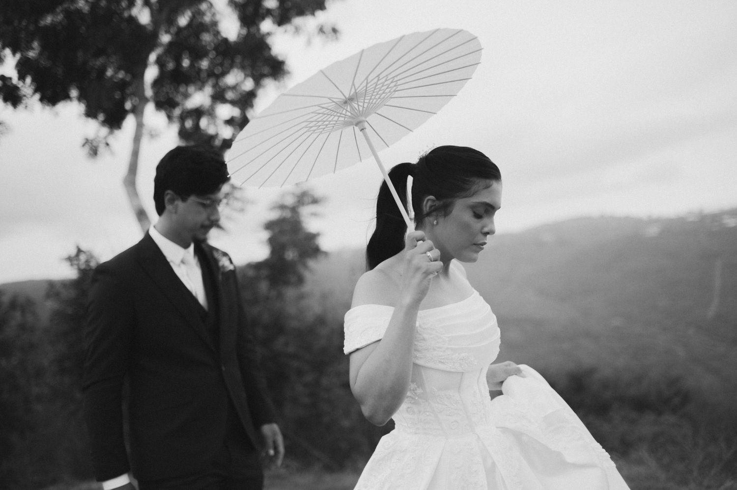 Black and white photo of a bride holding a white parasol and groom standing outdoors with lush hills in the background.
