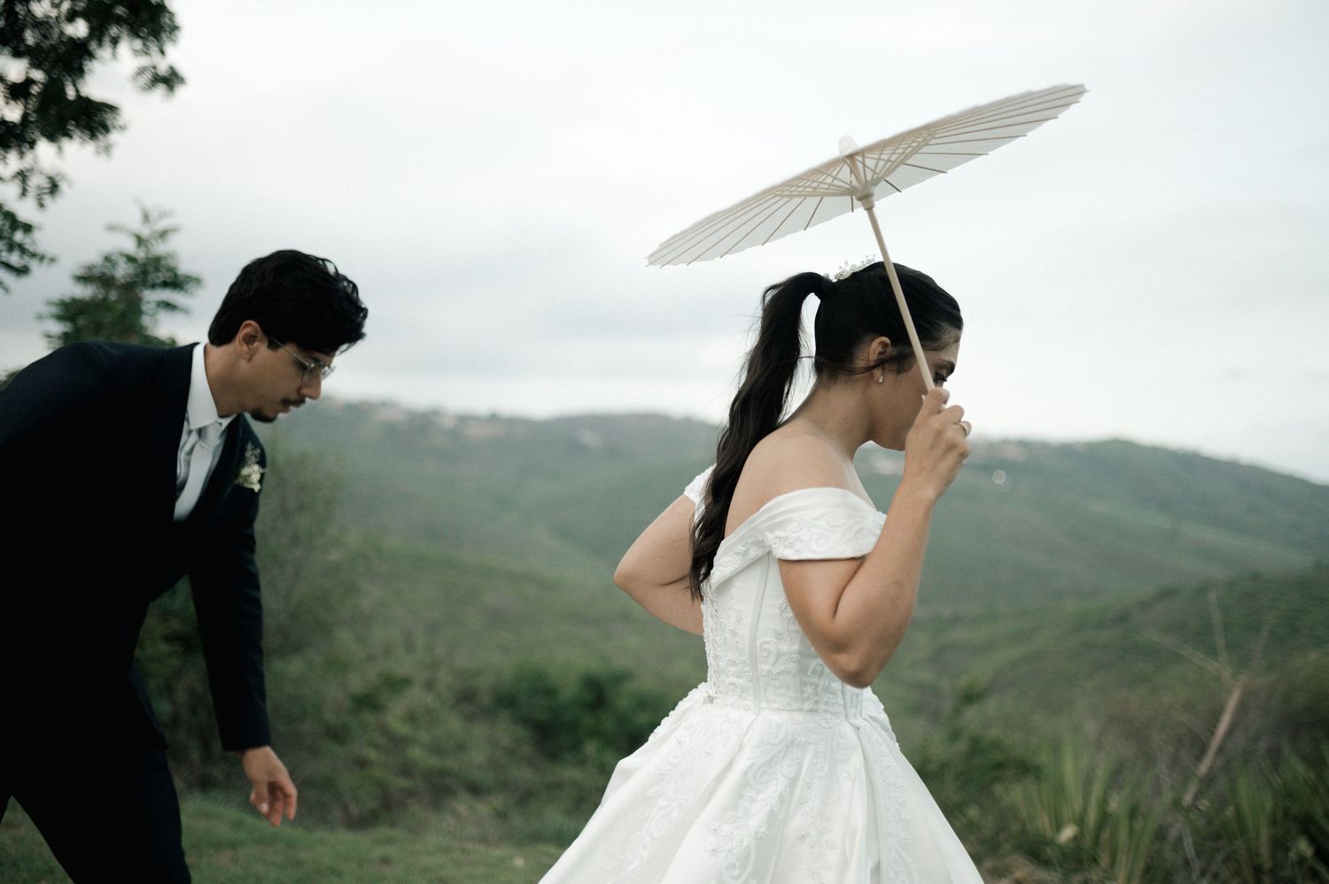 Bride in white off-shoulder gown holds parasol outdoors as groom adjusts attire, with lush green hills in background.