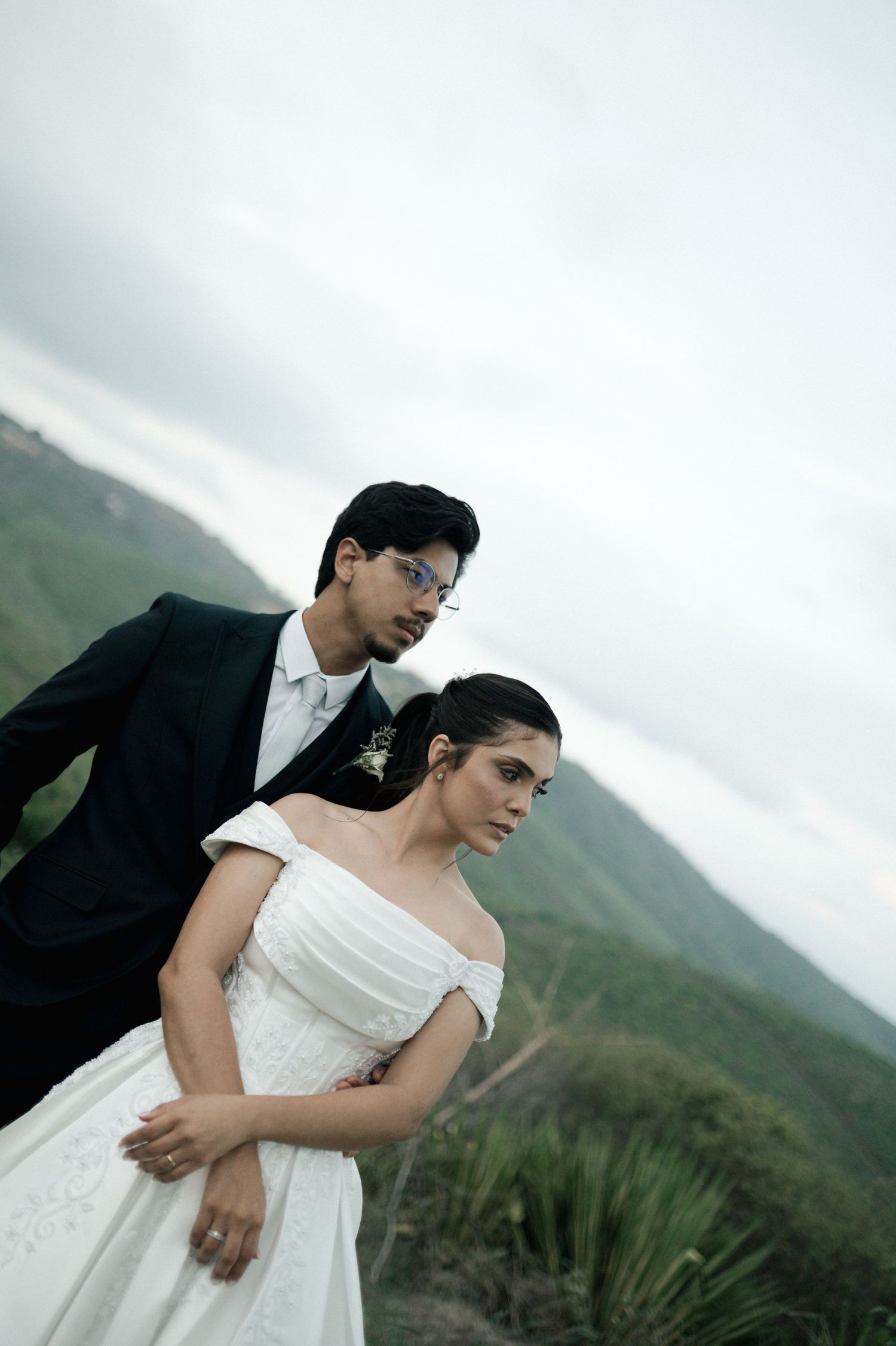 Couple in wedding attire posing on a scenic mountain overlook, bride in white off-shoulder gown, groom in black suit.