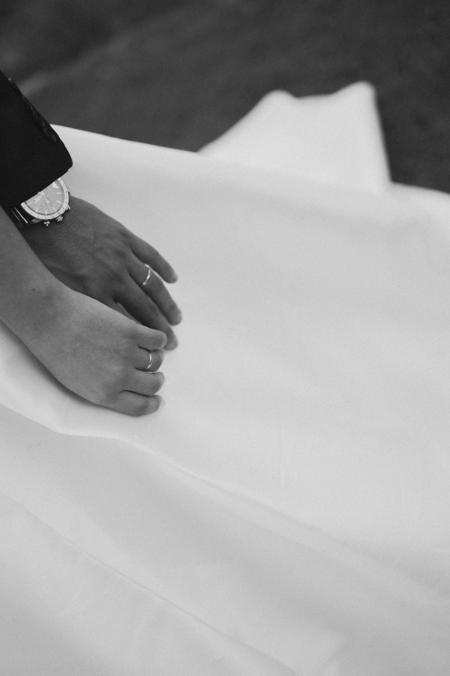 Black and white photo of a bride and groom holding hands, showing wedding rings and a watch on a white dress.