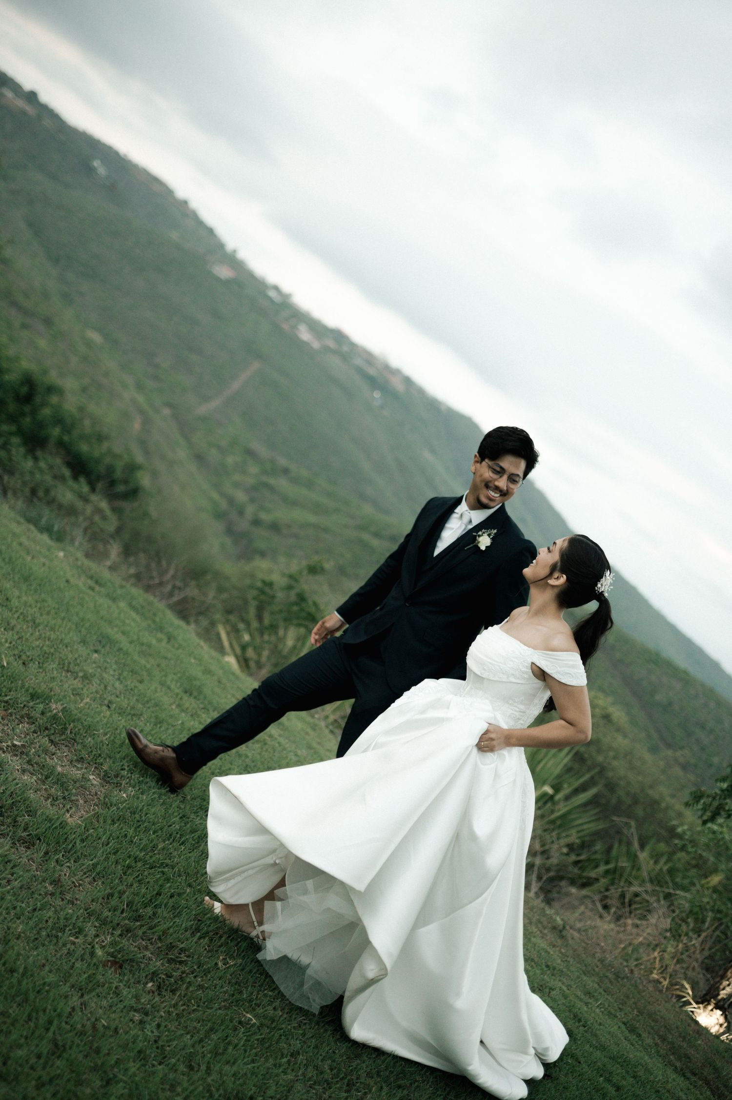 A joyful bride in a white gown lifts her groom in a black suit on a lush green hillside with misty mountains behind them.