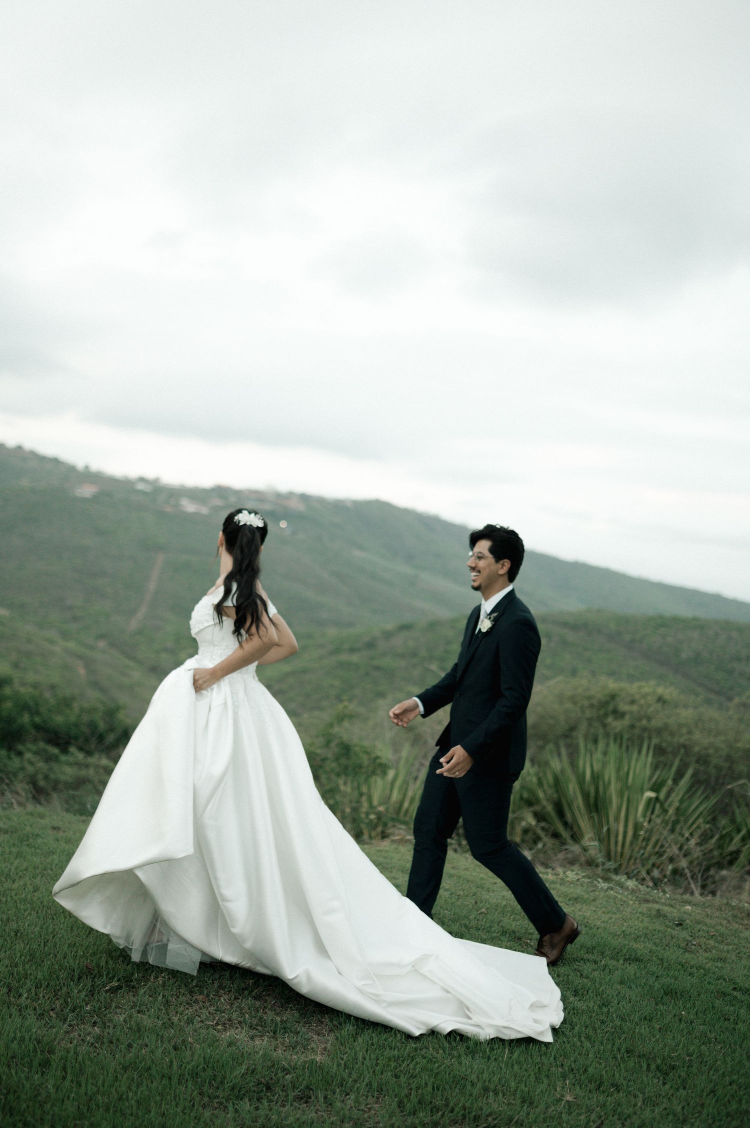 A bride in a flowing white gown and groom in a dark suit walk hand in hand on a green hillside.