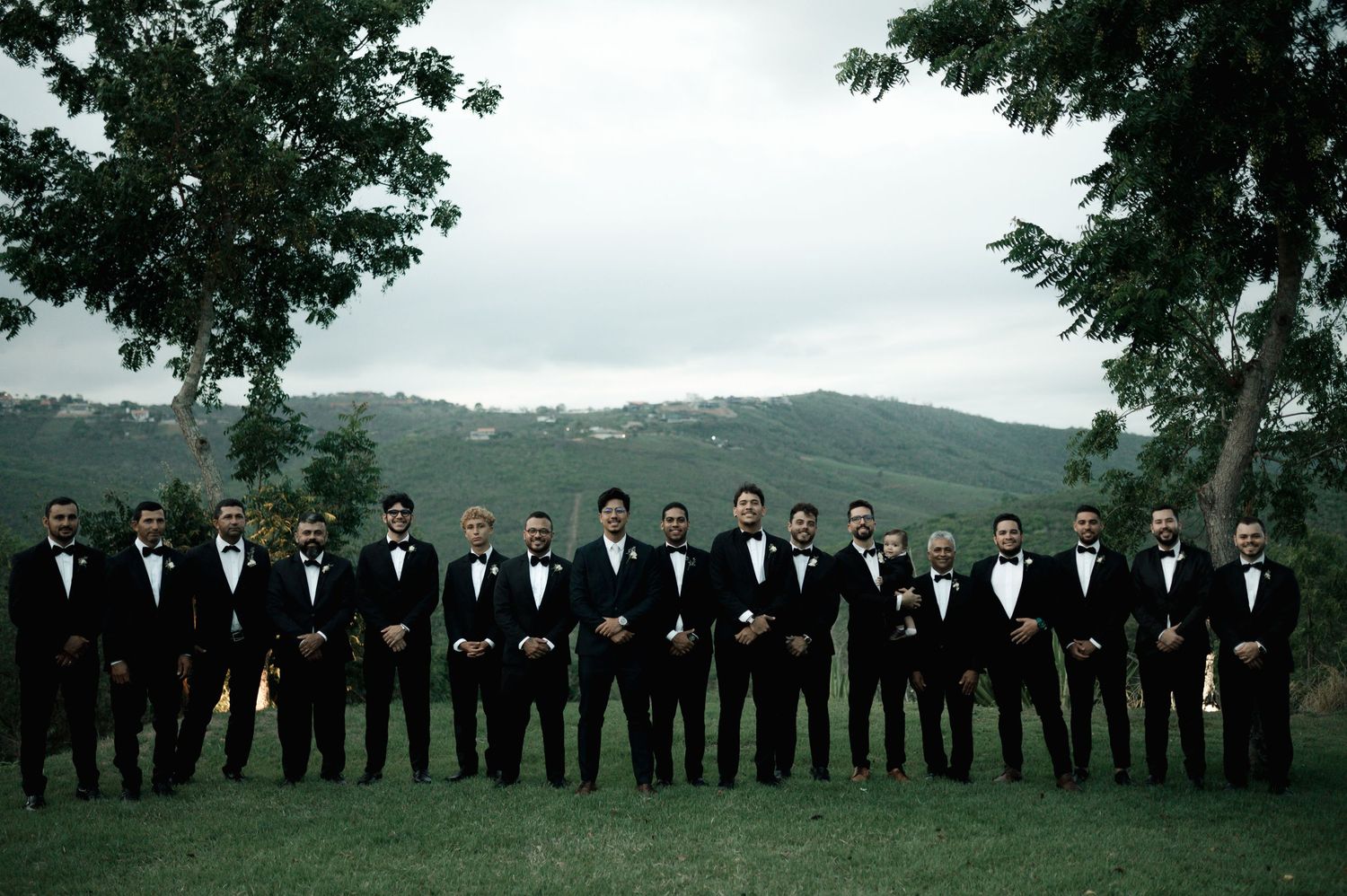 A groom and his groomsmen dressed in black tuxedos pose outdoors with rolling green hills in the background.