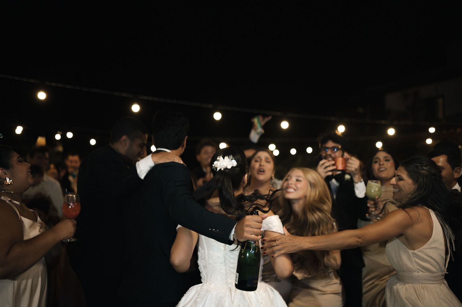 Bride and guests celebrating at a wedding reception at night, popping champagne under string lights.