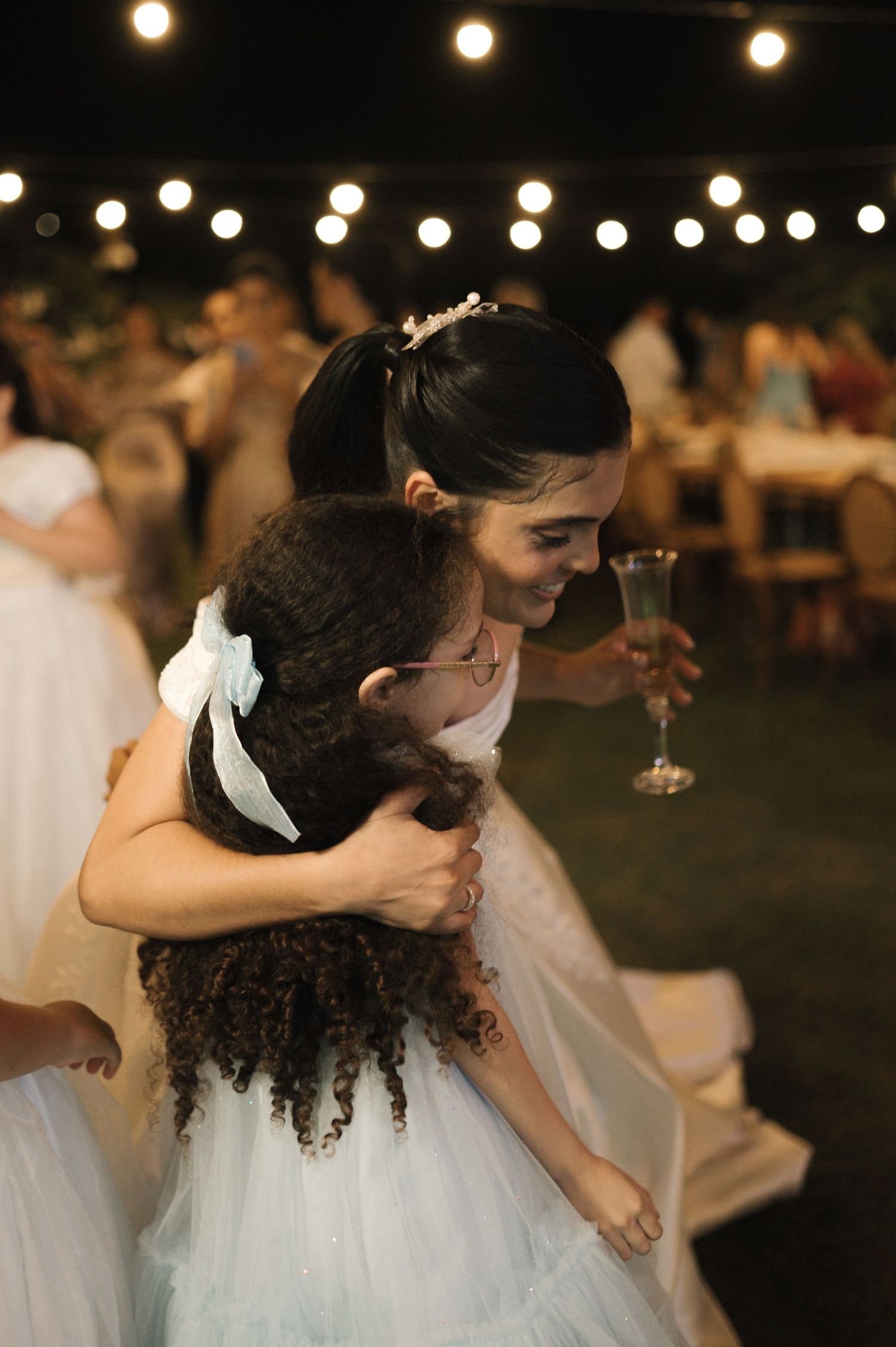 Bride in white gown hugging a young flower girl with curly hair at an outdoor wedding reception at night.