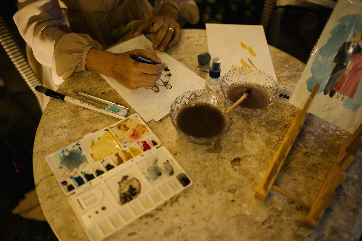 Artist painting with watercolors at a marble table, with brushes, paint palette, and water cup nearby.