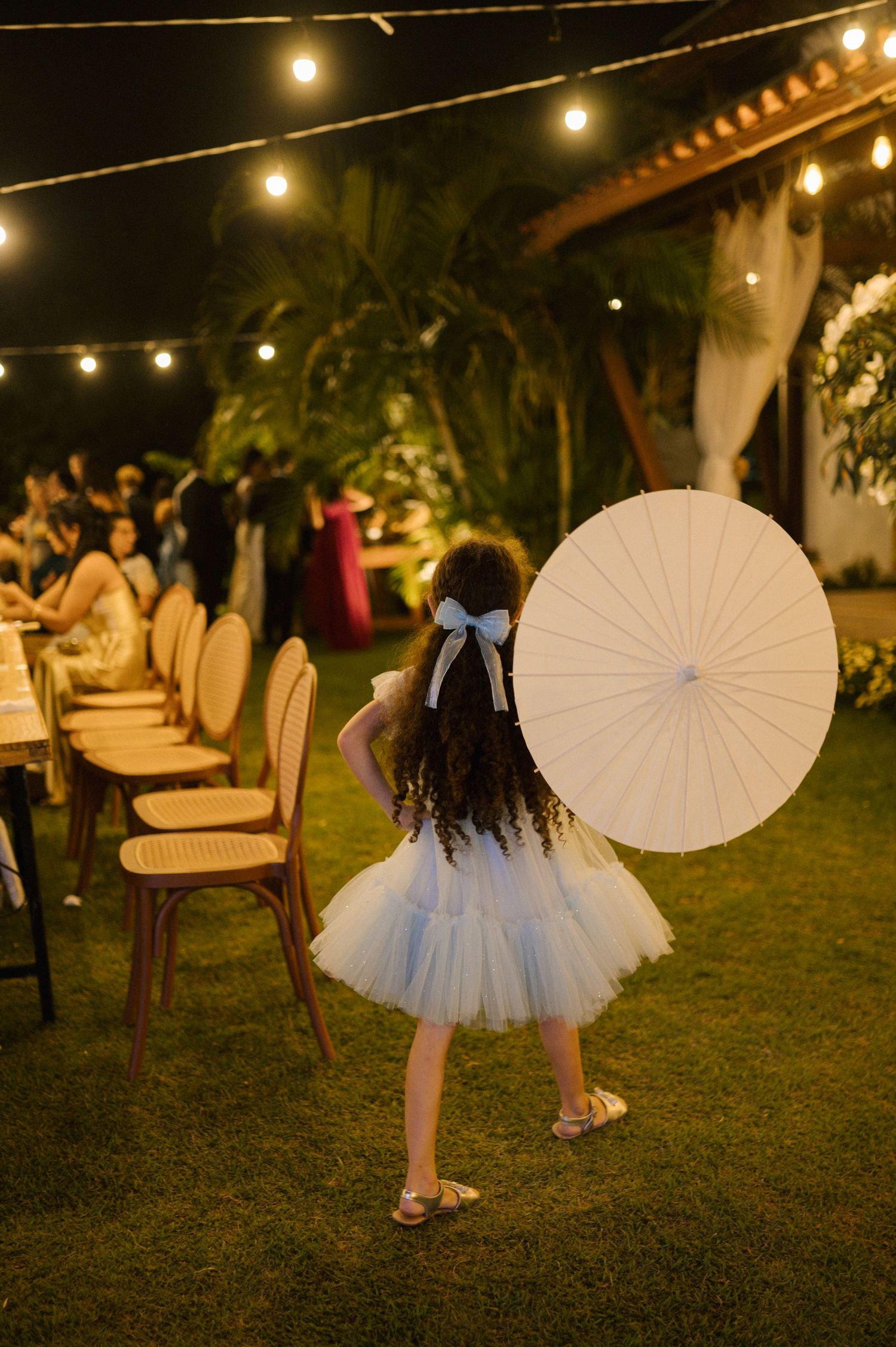 Young girl in white tutu dress holds parasol at elegant outdoor wedding reception with string lights at night.