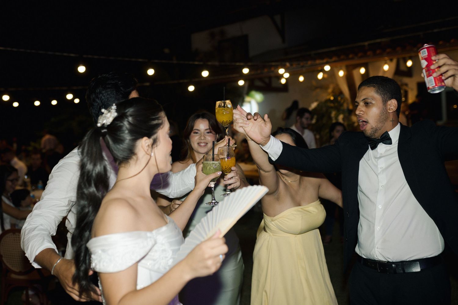Wedding guests toasting with drinks at an outdoor evening reception under string lights.