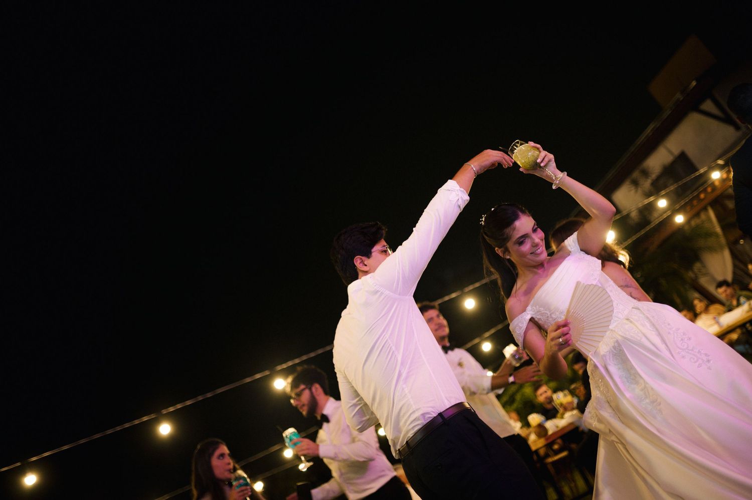 Couple sharing first dance at outdoor wedding reception under string lights at night.