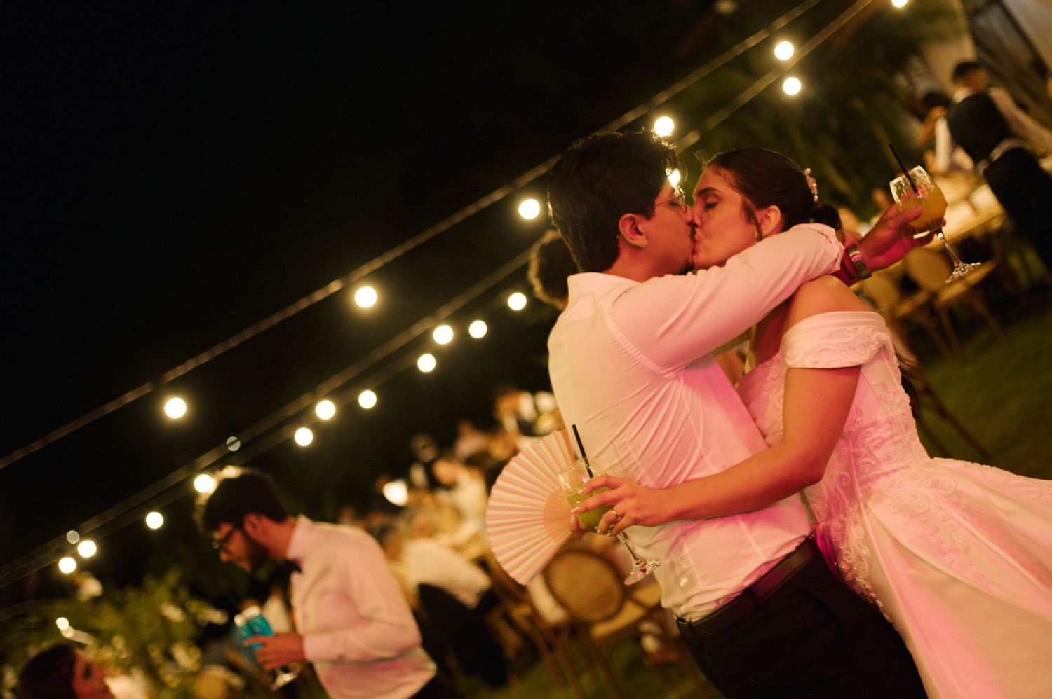 Couple sharing a romantic kiss at an outdoor evening wedding reception with string lights glowing in the background.