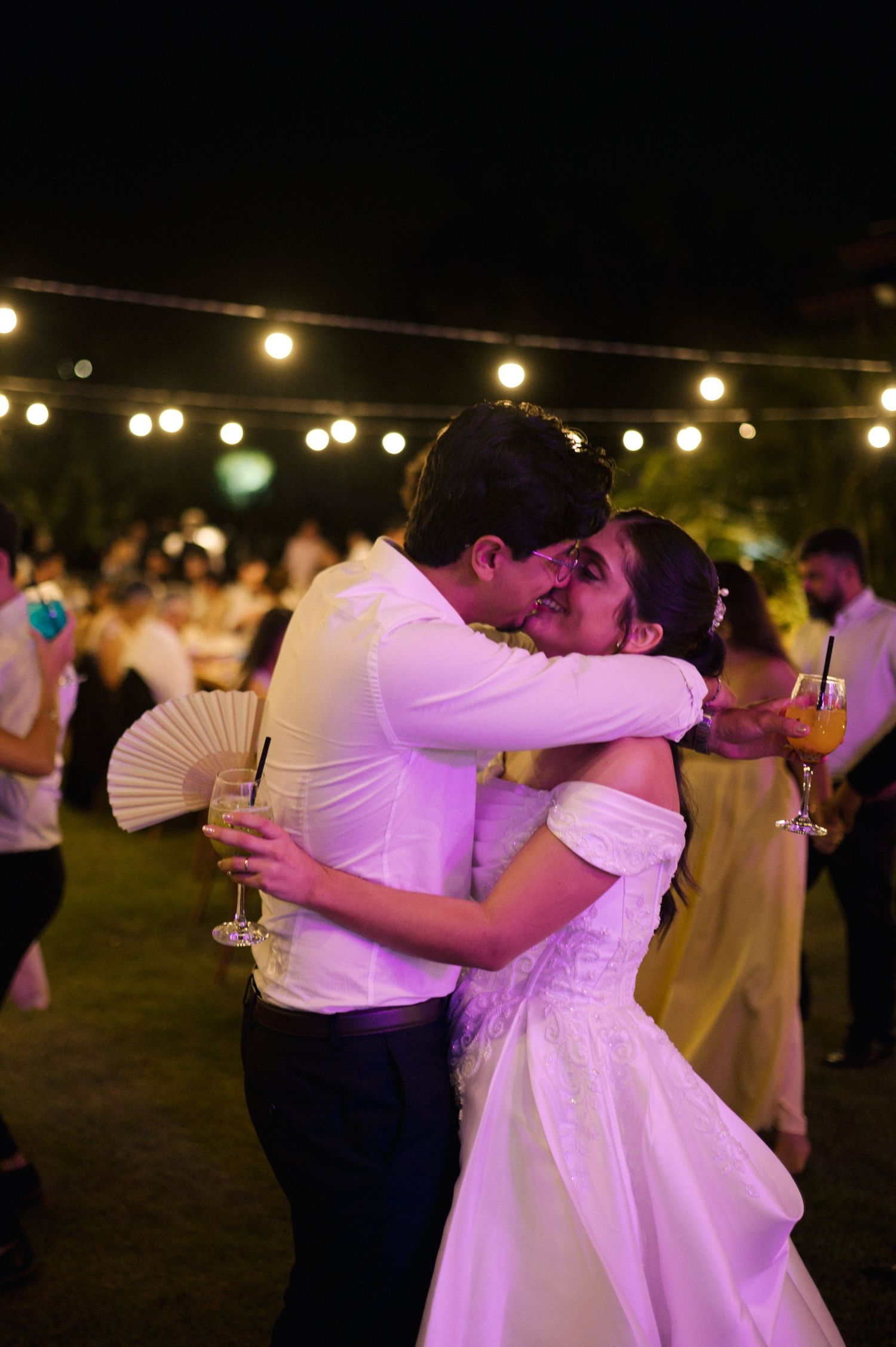 Bride and groom share a romantic kiss during their outdoor evening wedding reception under string lights.