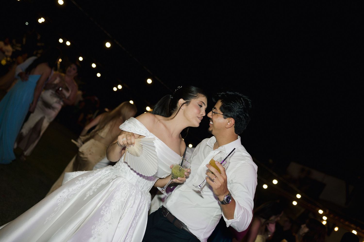 Couple sharing a romantic kiss in wedding attire at an outdoor evening reception with string lights.