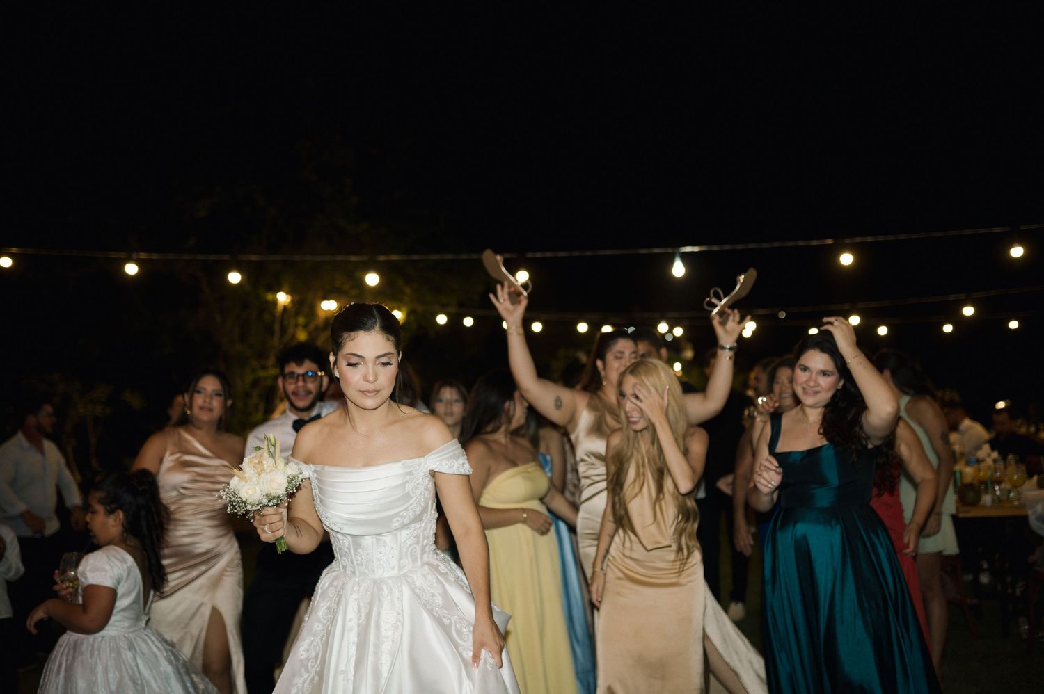 Bride in white off-shoulder gown holds bouquet while dancing with bridesmaids under string lights at outdoor wedding reception.