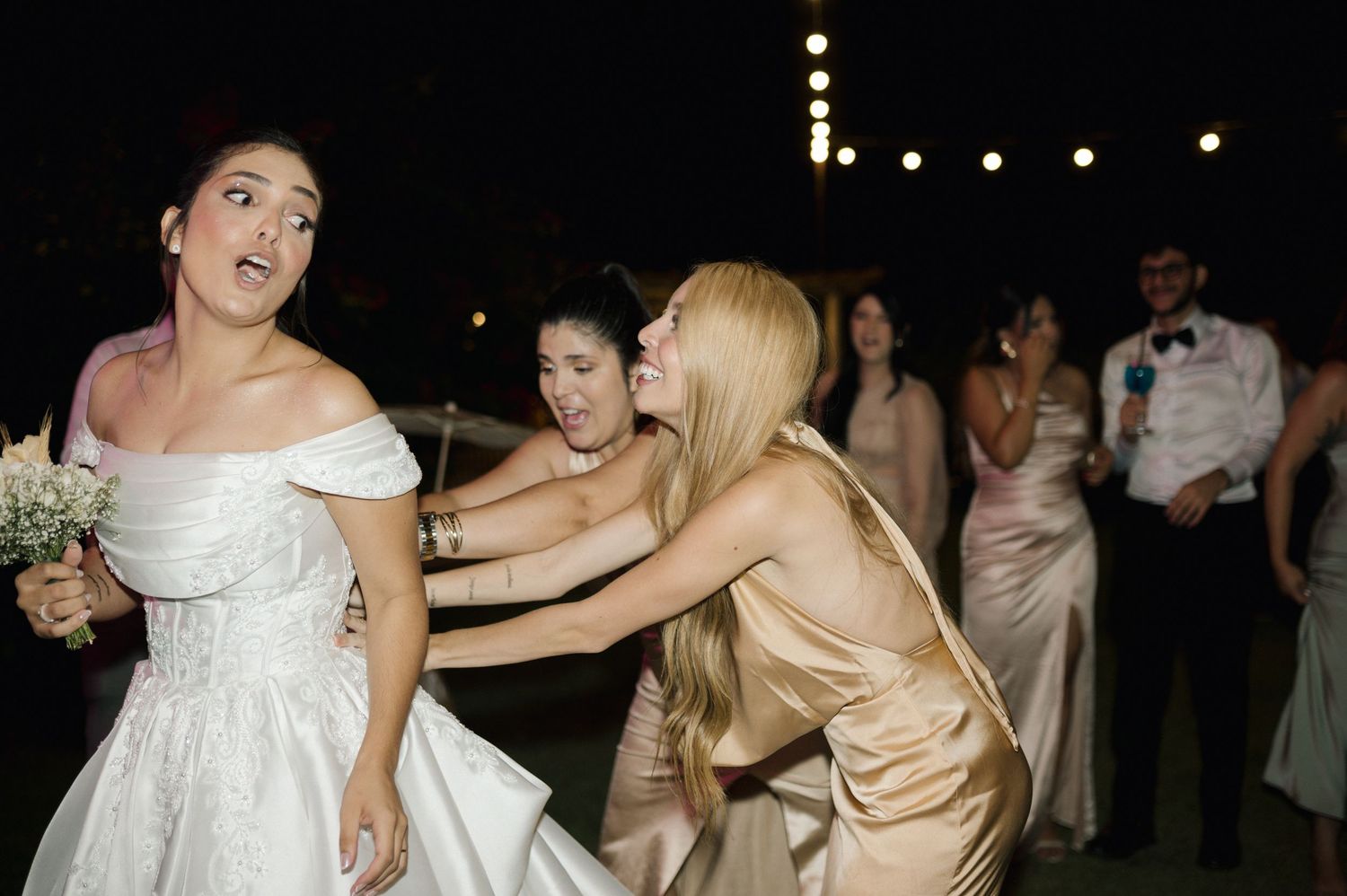 Bride in white off-shoulder gown reacts as bridesmaid in gold dress adjusts her outfit at outdoor evening wedding reception.