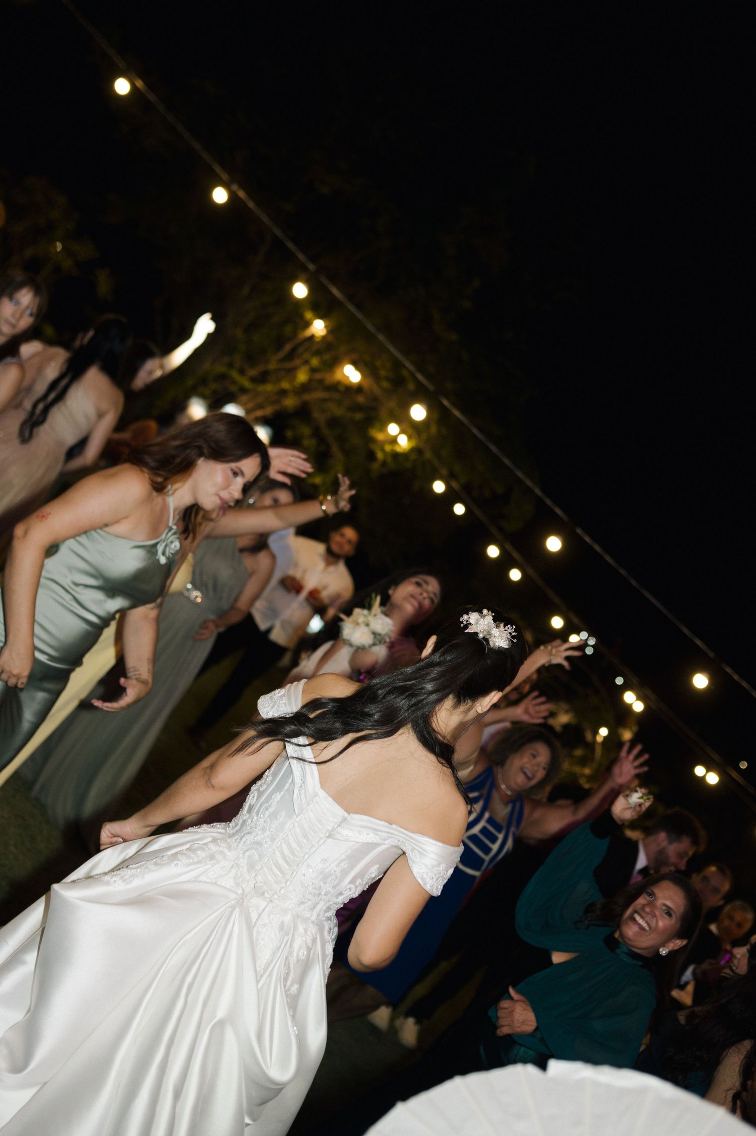 Bride tosses bouquet to excited guests at outdoor evening wedding reception with string lights.