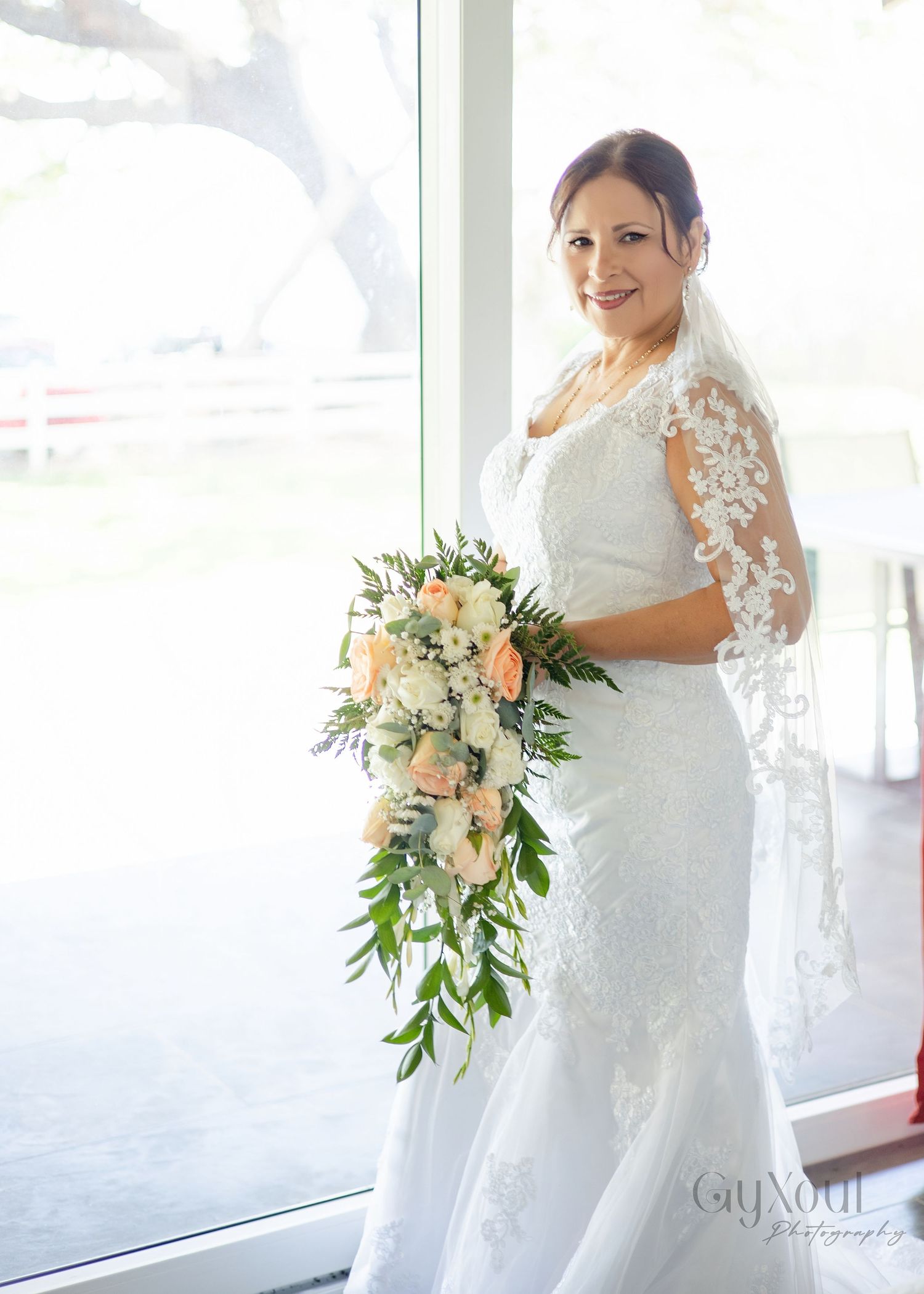 Bride in white lace gown and veil holding cascading peach and white floral bouquet standing by large window.