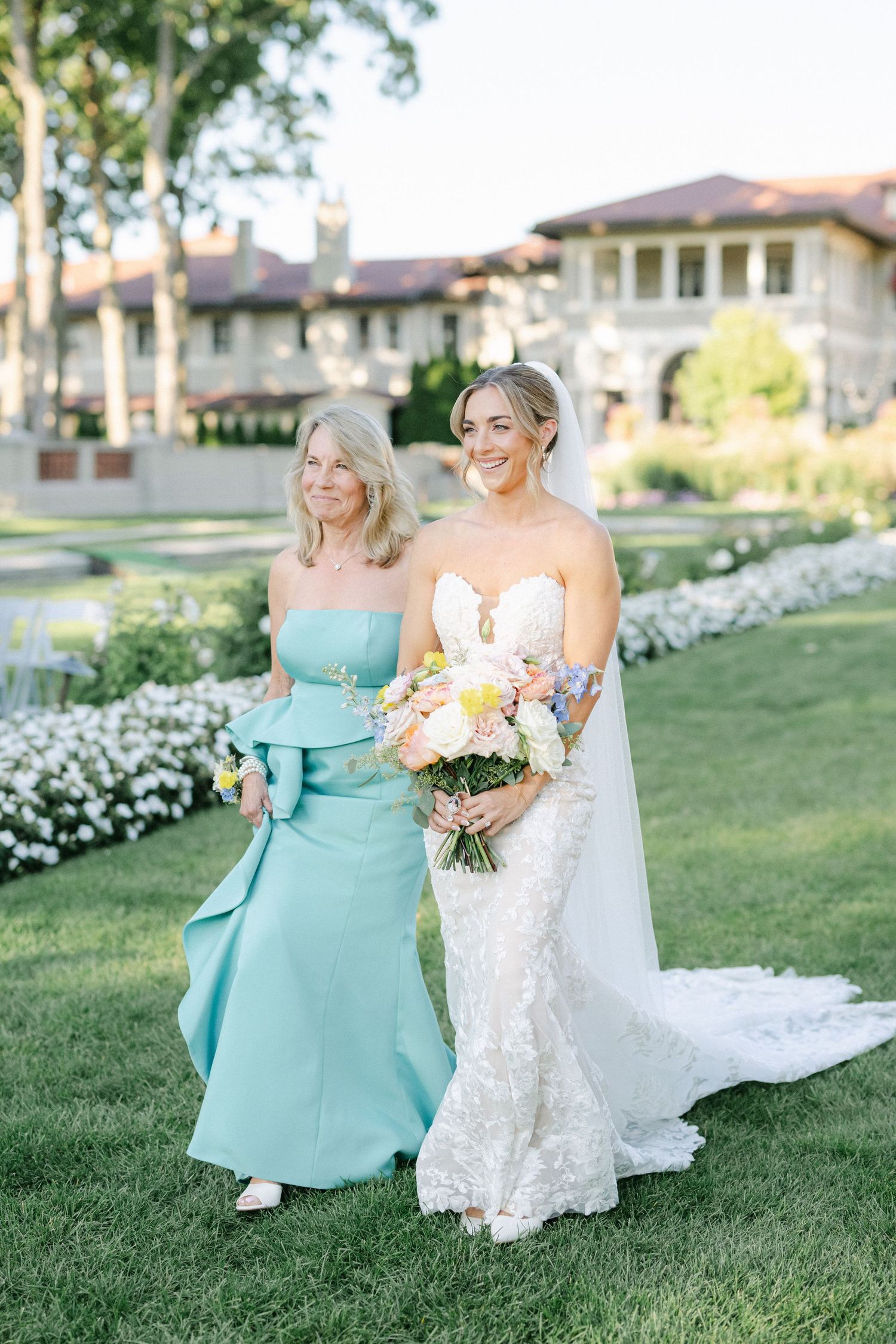 Armour House Wedding - Bride in lace gown holds floral bouquet walking with woman in teal dress on manicured lawn outside elegant venue.
