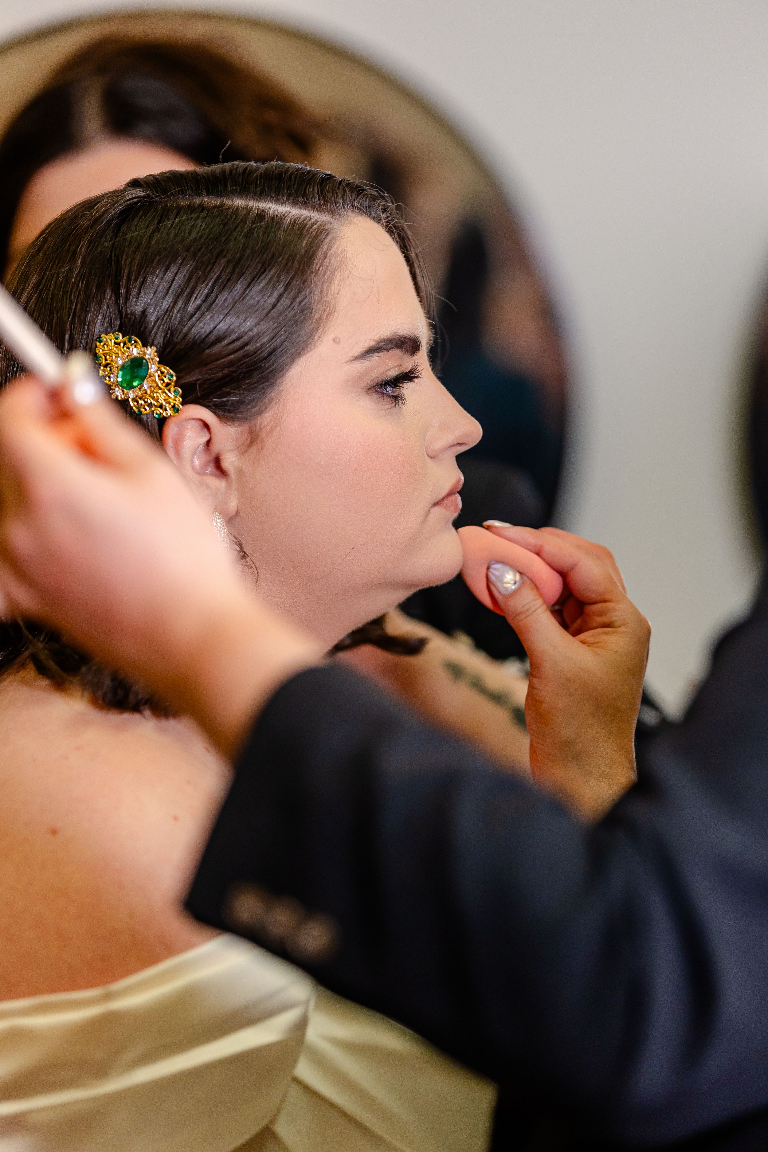 Bride gets makeup applied by artist, wearing gold and emerald hair accessory before her wedding ceremony.