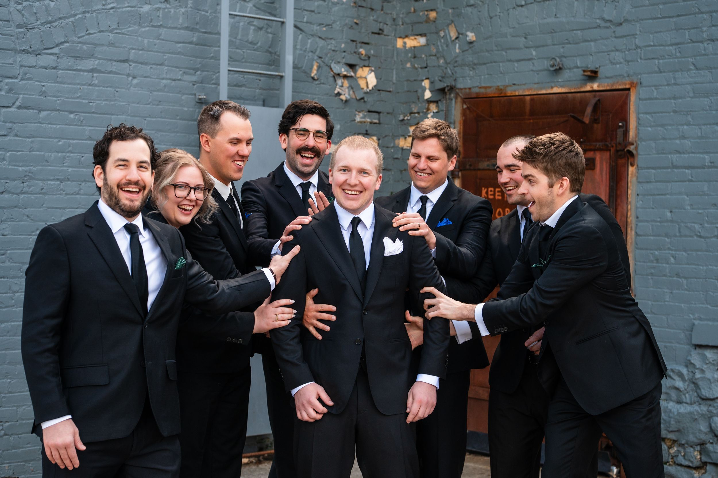 Groom in black suit smiles as groomsmen in dark suits playfully surround him outdoors against a gray brick wall.
