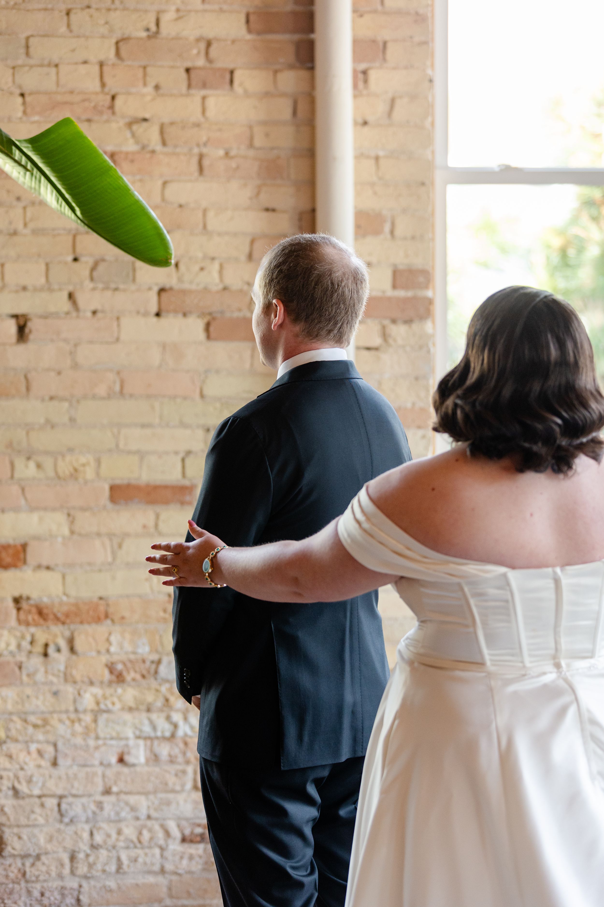 Bride in off-shoulder gown and groom in dark suit stand together facing a brick wall during wedding first look.