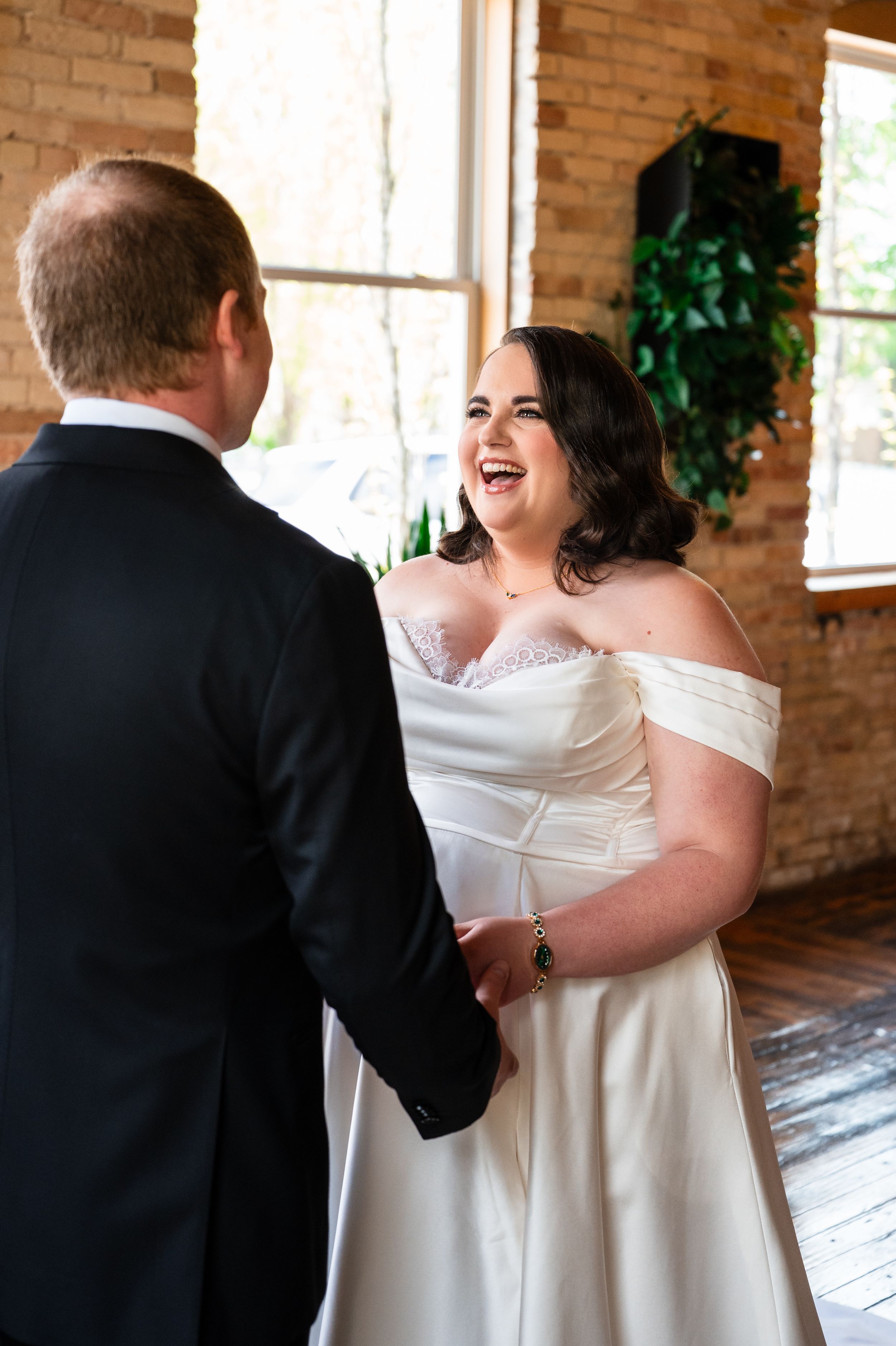 Bride in off-shoulder white gown laughs joyfully while holding hands with groom during wedding first look.