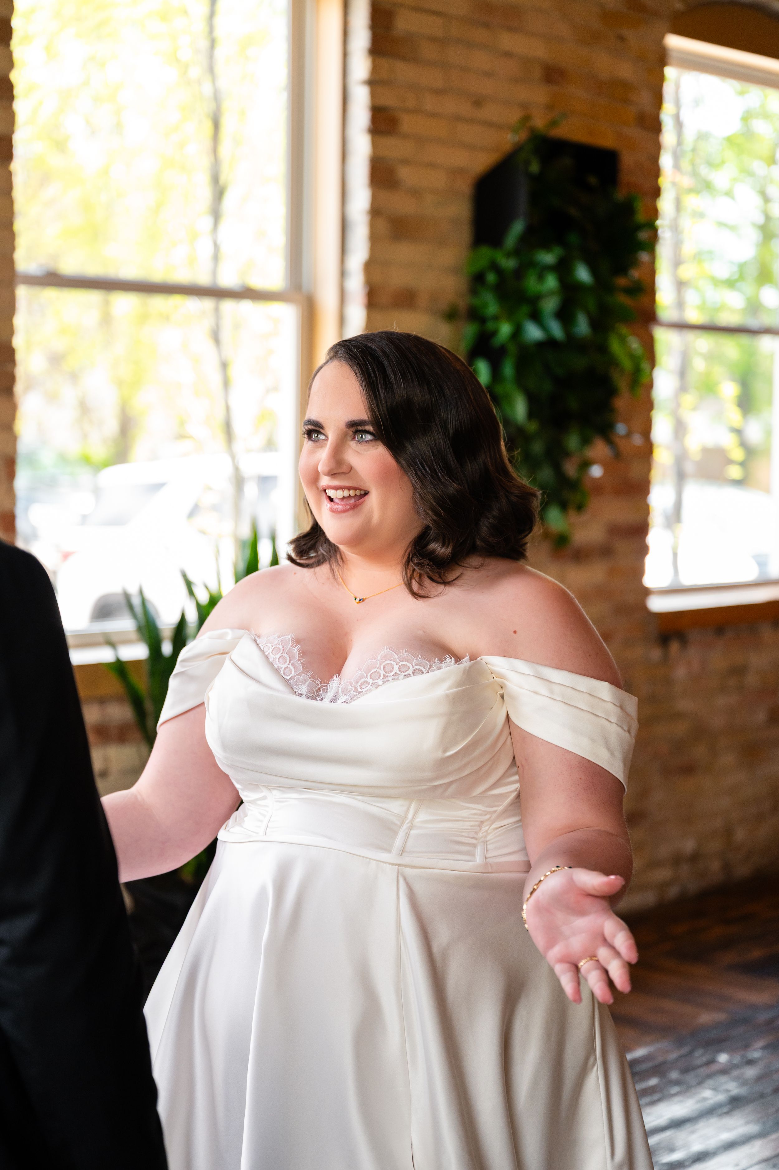 Smiling bride in an off-shoulder white satin wedding gown standing in a rustic brick-walled venue.