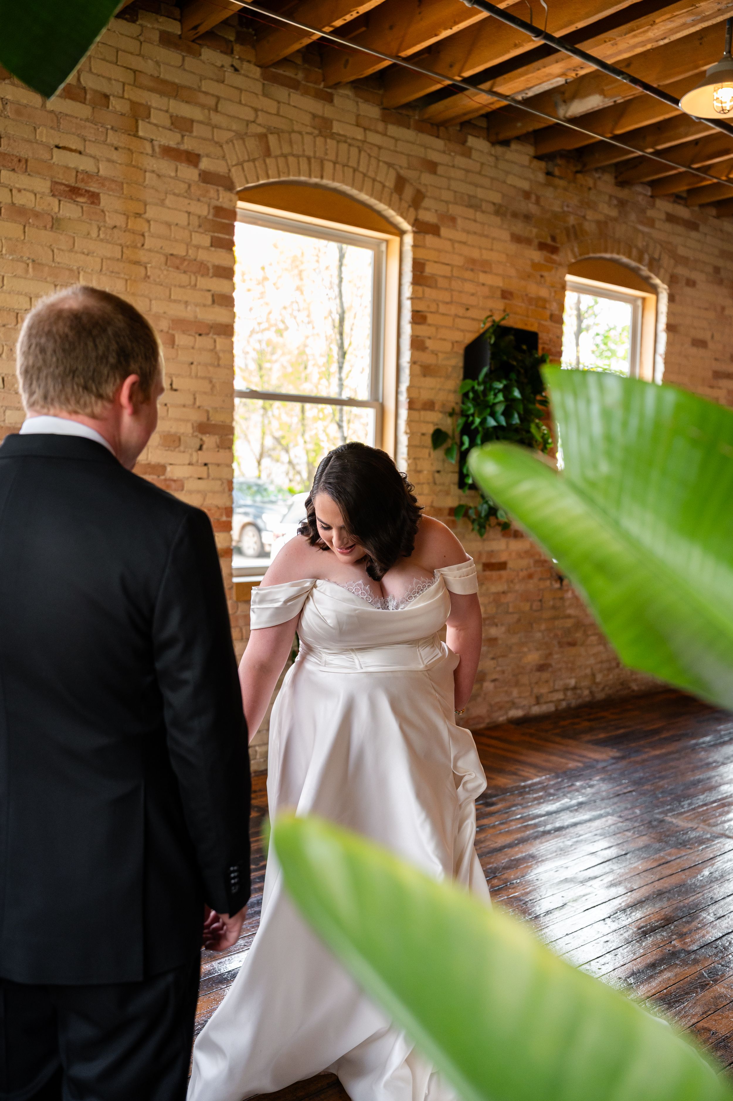 Bride in off-shoulder satin gown adjusting her dress beside groom in black suit in brick venue.