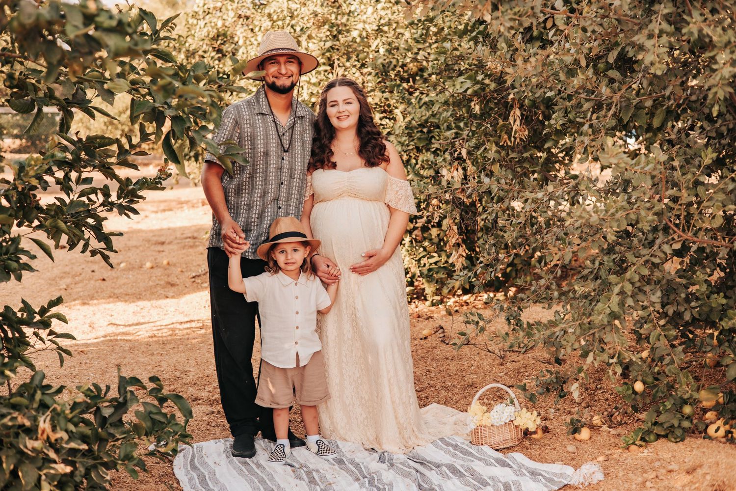 Pregnant woman in white gown poses with husband and toddler son in an apple orchard for a maternity photo.