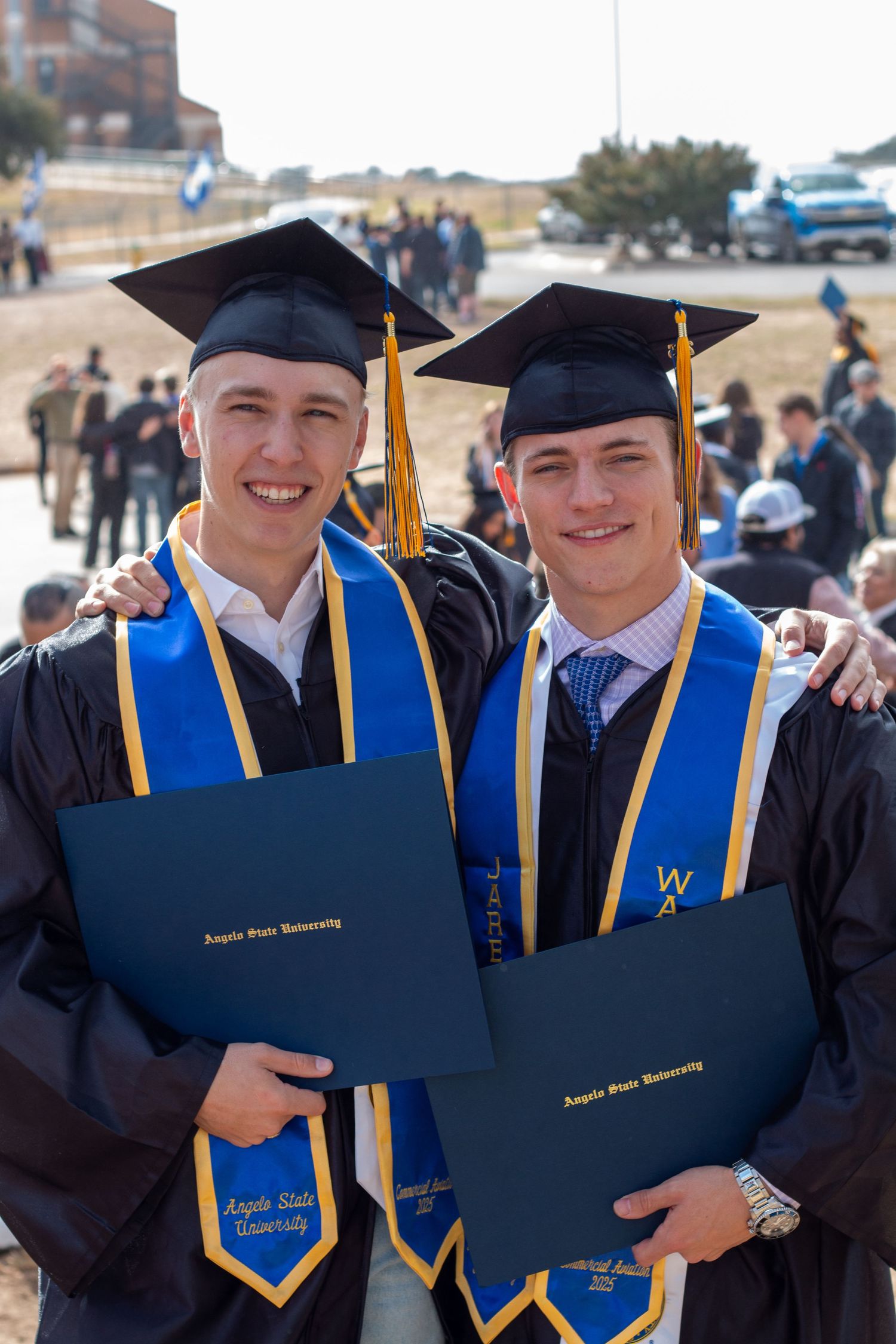 Two smiling male graduates in black caps and gowns with blue Angelo State University stoles holding diplomas outdoors.