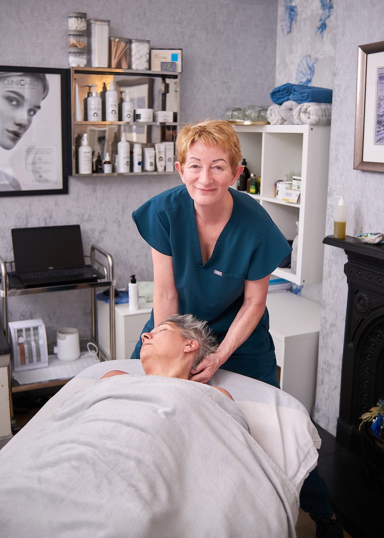 Esthetician in teal scrubs performing a relaxing facial massage on a client lying on a treatment table in a spa.