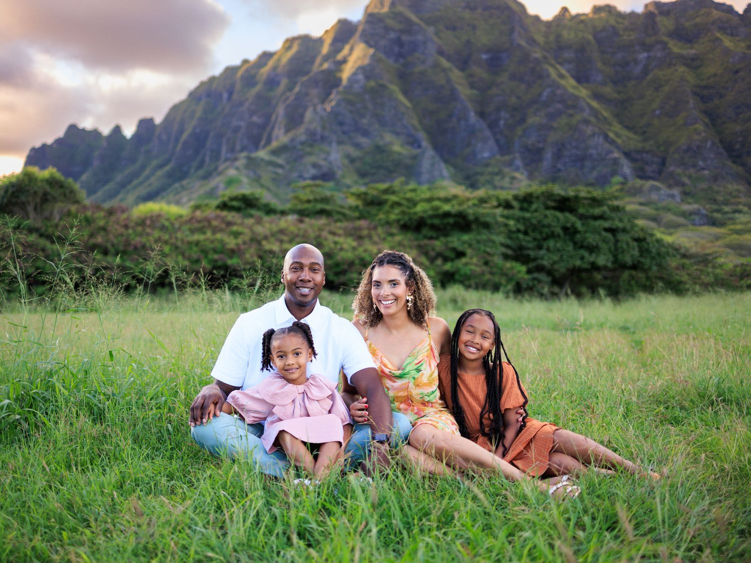 Family of four posing in lush green meadow with dramatic mountain backdrop at sunset in Hawaii.