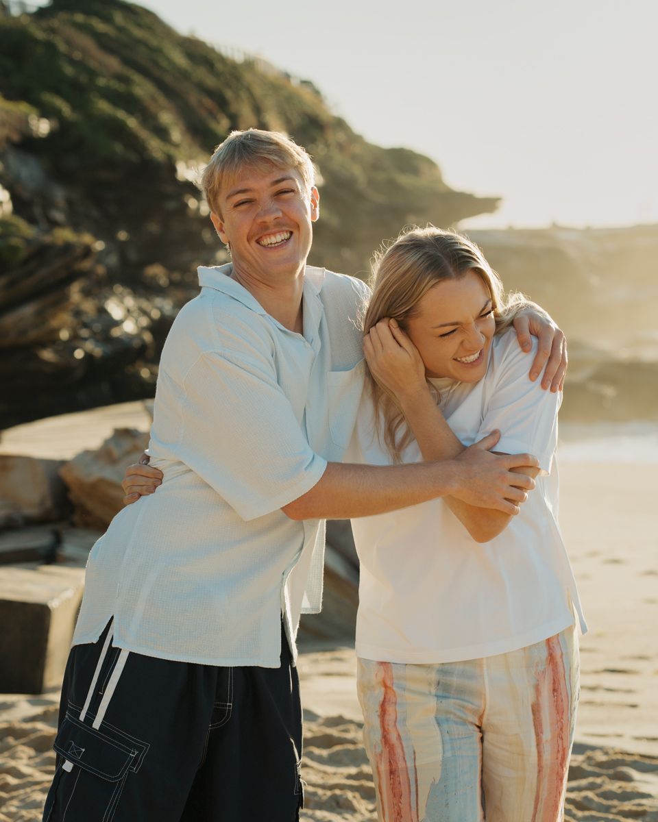 Two smiling adult siblings hugging on a sunny beach, wearing white tops, with rocky cliffs in the back at Bronte Beach family photoshoot