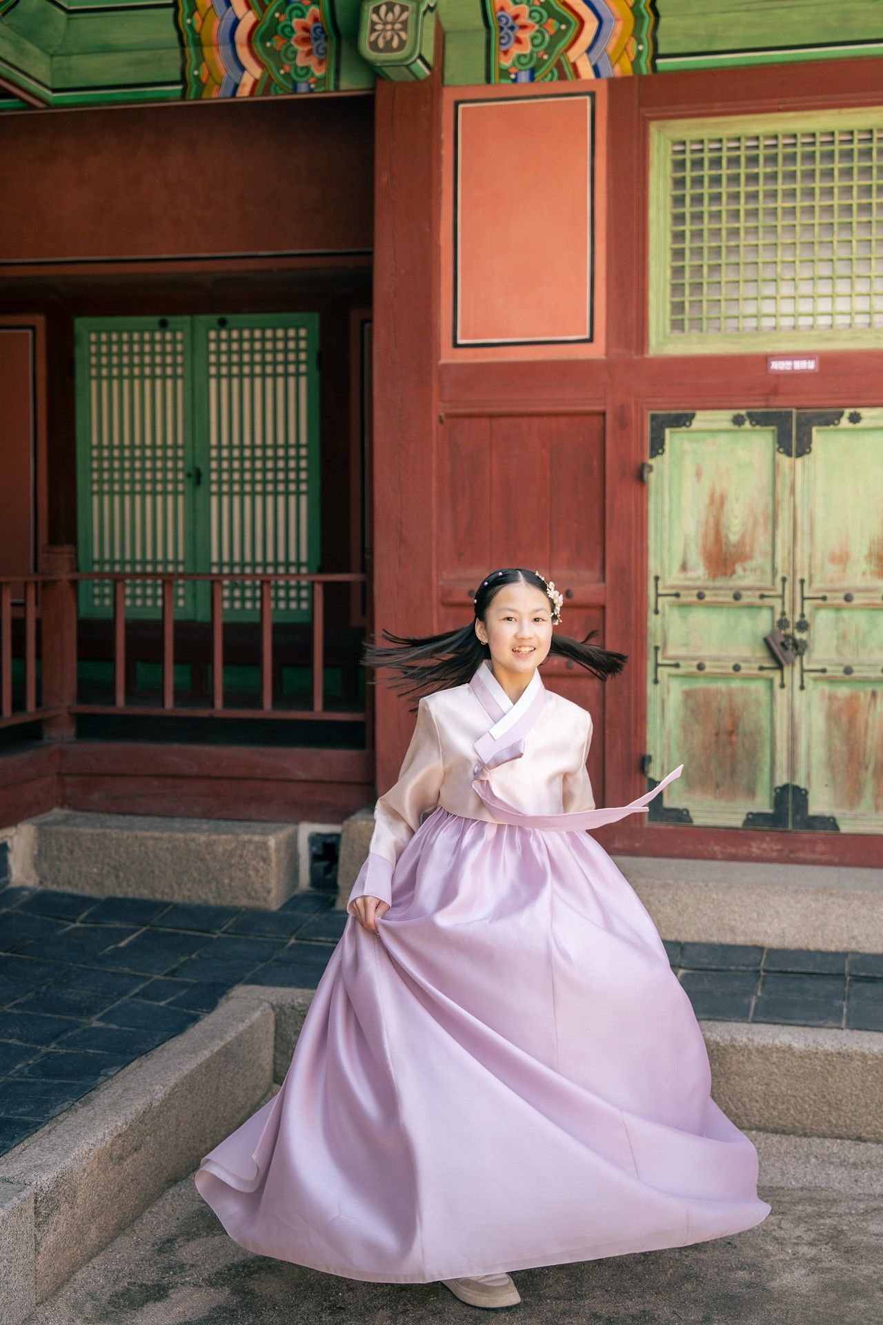 Young woman twirling in pink hanbok traditional Korean dress at historic palace with red and green architecture.