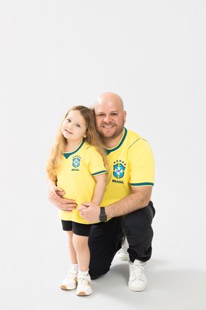 Bald man in Brazil jersey crouching and hugging young blonde girl in matching yellow Brasil shirt.
