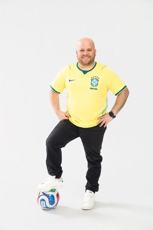 Bald man in Brazil yellow Nike soccer jersey posing with foot on soccer ball against white background.