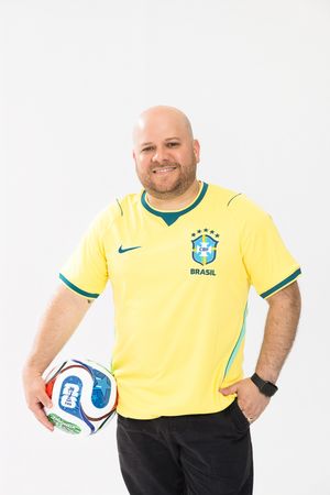 Smiling bald man wearing a yellow Brazil CBF Nike soccer jersey holding a football against a white background.