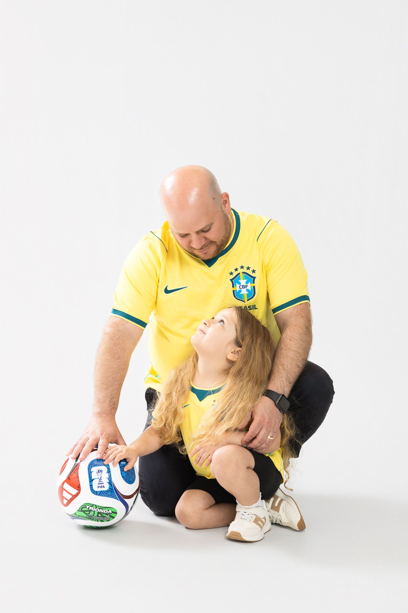 Bald man in Brazil jersey sitting with young girl in matching shirt while touching a soccer ball.