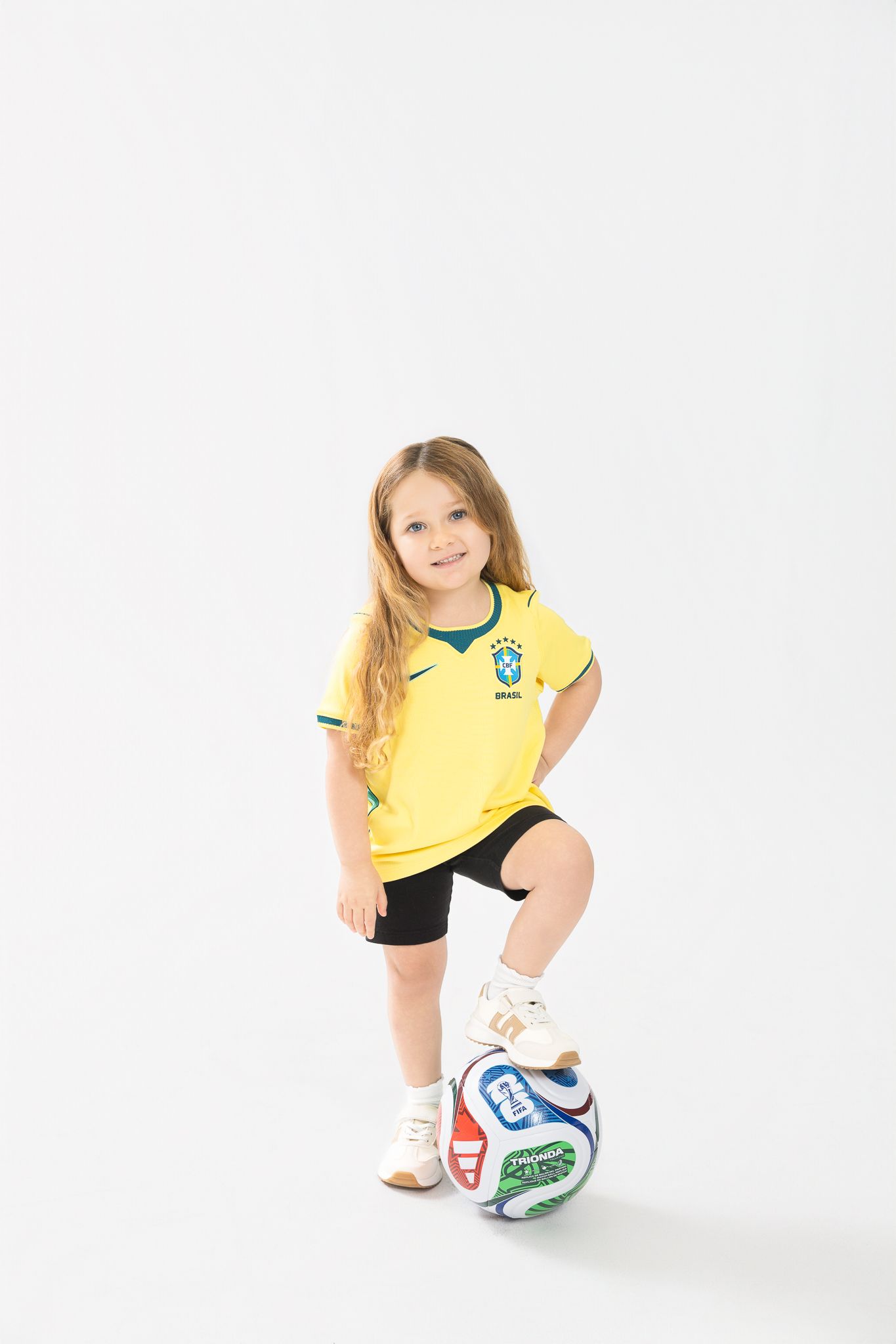 Young girl in Brazil soccer jersey with foot on soccer ball, posing in studio on white background.