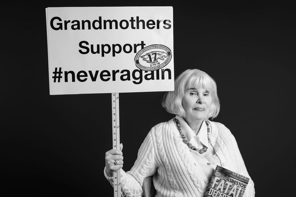 Elderly woman holding a sign reading Grandmothers Support #neveragain in a black and white portrait photo.