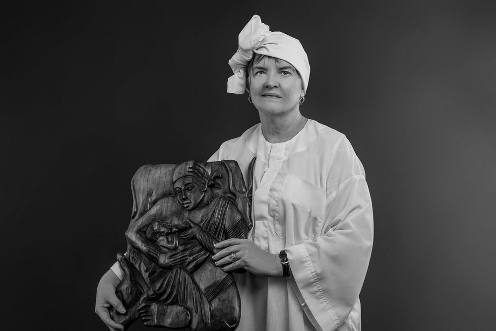 Black and white portrait of a woman in white attire holding a carved wooden sculpture, looking upward.