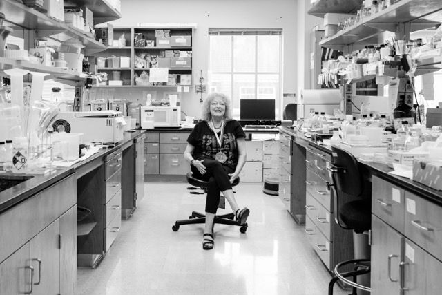 Woman scientist smiling in a research laboratory surrounded by equipment and shelves.
