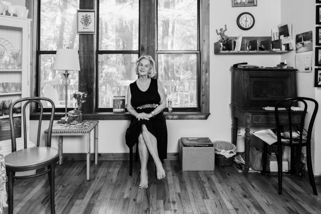 Woman in black dress sits by large windows in a rustic room with wooden floors and vintage furniture.