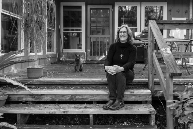 Woman smiling on wooden deck steps outside home, with cat visible in background amid lush garden.